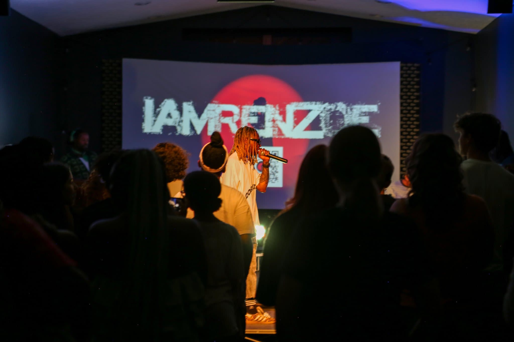 Renzoe performs in front of a screen reading IAMRENZOE, the crowd in silhouette, at Summer Glory.